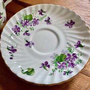 K&ouml;nnte beinhalten: Wei&szlig;e Untertasse mit gewelltem Rand und goldenem Rand. Die Untertasse ist mit lila Veilchen und gr&uuml;nen Bl&auml;ttern verziert. Das Design wiederholt sich am Rand der Untertasse. Eine passende Teetasse ist teilweise sichtbar.