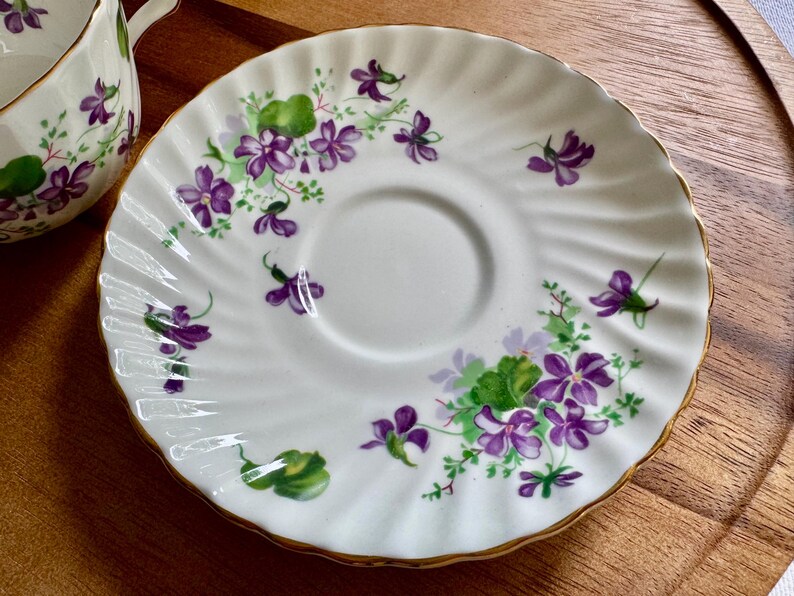 K&ouml;nnte beinhalten: Wei&szlig;e Untertasse mit gewelltem Rand und goldenem Rand. Die Untertasse ist mit lila Veilchen und gr&uuml;nen Bl&auml;ttern verziert. Das Design wiederholt sich am Rand der Untertasse. Eine passende Teetasse ist teilweise sichtbar.