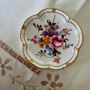May include: A white, scalloped-edge dish with a floral design. The dish features a gold rim and a bouquet of pink, purple, yellow, and blue flowers in the center. The dish is likely made of porcelain or ceramic.