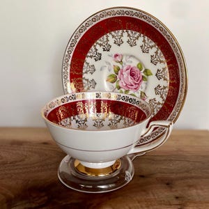 Pode incluir: Conjunto de ch&aacute; vintage branco com design vermelho e dourado. A ch&aacute;vena e o pires apresentam um motivo de rosa rosa. O conjunto &eacute; exibido em um suporte de vidro transparente, para uma est&eacute;tica elegante.