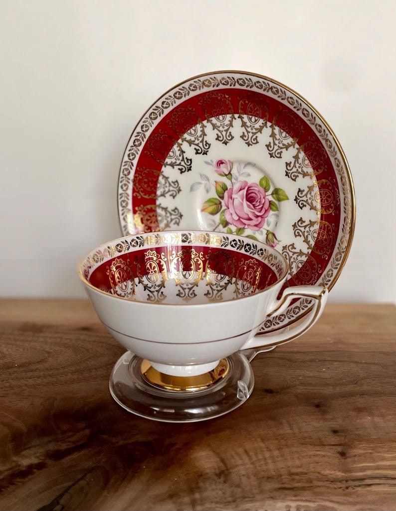 Pode incluir: Conjunto de ch&aacute; vintage branco com design vermelho e dourado. A ch&aacute;vena e o pires apresentam um motivo de rosa rosa. O conjunto &eacute; exibido em um suporte de vidro transparente, para uma est&eacute;tica elegante.