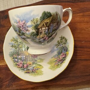 May include: A white teacup and saucer with a floral pattern featuring a cottage in a countryside setting. The cup and saucer have a gold trim.