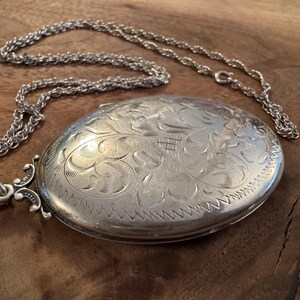 Puede incluir: Un collar con un medall&oacute;n ovalado plateado con un intrincado dise&ntilde;o floral en la parte delantera. El medall&oacute;n est&aacute; sujeto a una delicada cadena. El fondo es una superficie de madera.