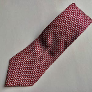 Puede incluir: Una corbata con un fondo rojo intenso y un patr&oacute;n repetitivo de peque&ntilde;as formas de tr&eacute;bol de cuatro hojas en color blanco. La corbata tiene una punta afilada y una textura tejida suave, mostrando un dise&ntilde;o geom&eacute;trico consistente.