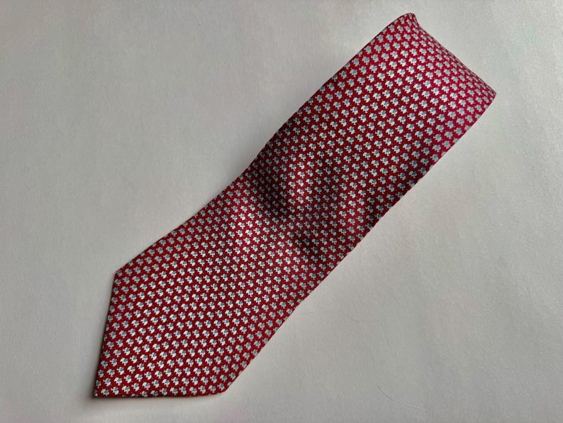 Puede incluir: Una corbata con un fondo rojo intenso y un patr&oacute;n repetitivo de peque&ntilde;as formas de tr&eacute;bol de cuatro hojas en color blanco. La corbata tiene una punta afilada y una textura tejida suave, mostrando un dise&ntilde;o geom&eacute;trico consistente.