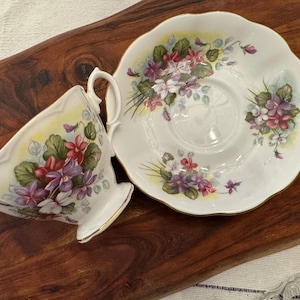 May include: A white teacup and saucer with a floral design featuring purple, pink, and white flowers. The flowers are painted in a realistic style and are surrounded by green leaves. The teacup and saucer have a gold trim.