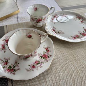 May include: Two white teacups with floral patterns and gold trim, one on a matching saucer with a floral pattern and gold trim, and the other on a larger white plate with a floral pattern and gold trim.