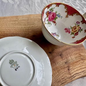 May include: A white teacup and saucer set with floral designs and gold trim. The teacup features a red patterned border and a handle. The saucer is white with a scalloped edge. The set is on a wooden surface.
