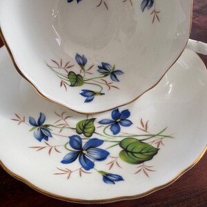 May include: A white teacup and saucer set with a gold rim and a floral design featuring blue and green violets.
