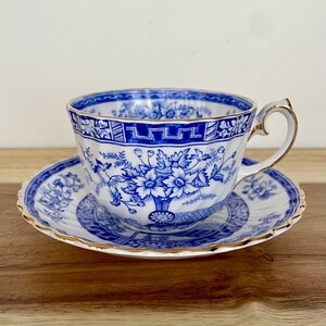 May include: A white teacup and saucer set with a blue floral design. The teacup has a gold handle and trim. The saucer has a scalloped edge. The set is on a wooden surface.