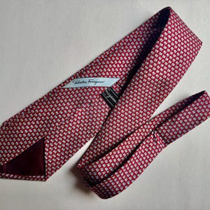 Puede incluir: Una corbata roja con un patr&oacute;n repetitivo de peque&ntilde;os motivos florales blancos y azul claro. La corbata incluye una etiqueta de tela blanca con el texto Salvatore Ferragamo y una etiqueta de marca negra en el forro interior.