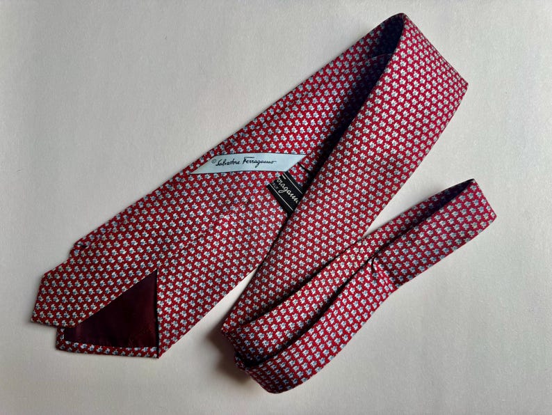 Puede incluir: Una corbata roja con un patr&oacute;n repetitivo de peque&ntilde;os motivos florales blancos y azul claro. La corbata incluye una etiqueta de tela blanca con el texto Salvatore Ferragamo y una etiqueta de marca negra en el forro interior.