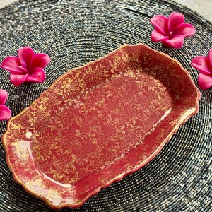 May include: A red and gold ceramic serving tray with a floral pattern. The tray is on a black beaded surface with pink flowers.