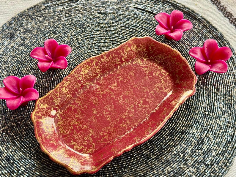 May include: A red and gold ceramic serving tray with a floral pattern. The tray is on a black beaded surface with pink flowers.