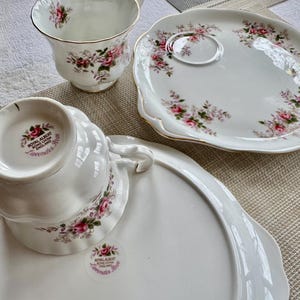 May include: A white teacup and saucer set with a floral pattern. The teacup and saucer are both decorated with pink and purple flowers. The saucer has a scalloped edge. The teacup has the words "Royal Albert Bone China England Lavender Rose" on the bottom.