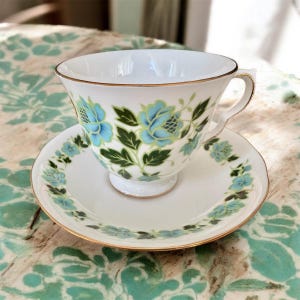 May include: A white teacup and saucer with a blue and green floral pattern. The cup and saucer have a gold trim.
