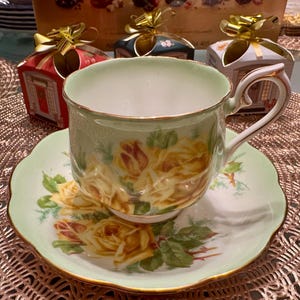 May include: A green and white teacup and saucer set with a floral design featuring yellow roses. The cup has a gold rim and the saucer has a scalloped edge.