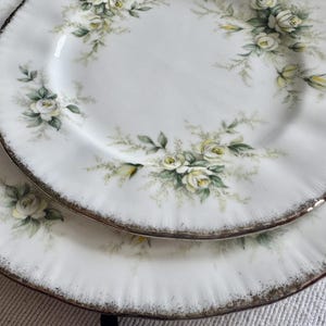 May include: Two white ceramic plates with a gold rim and a floral design featuring white roses and green leaves.