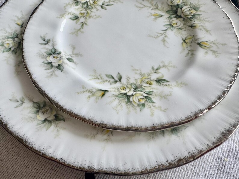 May include: Two white ceramic plates with a gold rim and a floral design featuring white roses and green leaves.