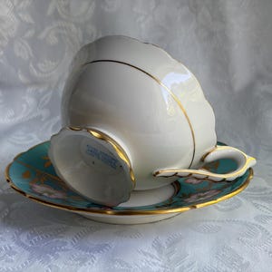 May include: A white porcelain teacup and saucer set featuring a scalloped edge and gold trim. The saucer has a teal border with floral accents. The underside of the teacup base displays blue text reading Royal Stafford, Made in England.