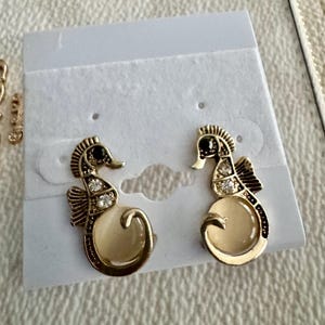 May include: A pair of gold-toned seahorse earrings with black accents and clear crystal embellishments. The seahorses have a cream-colored belly and are displayed on a white earring card.