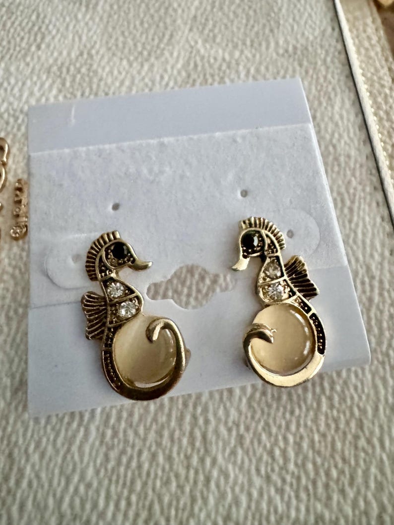 May include: A pair of gold-toned seahorse earrings with black accents and clear crystal embellishments. The seahorses have a cream-colored belly and are displayed on a white earring card.
