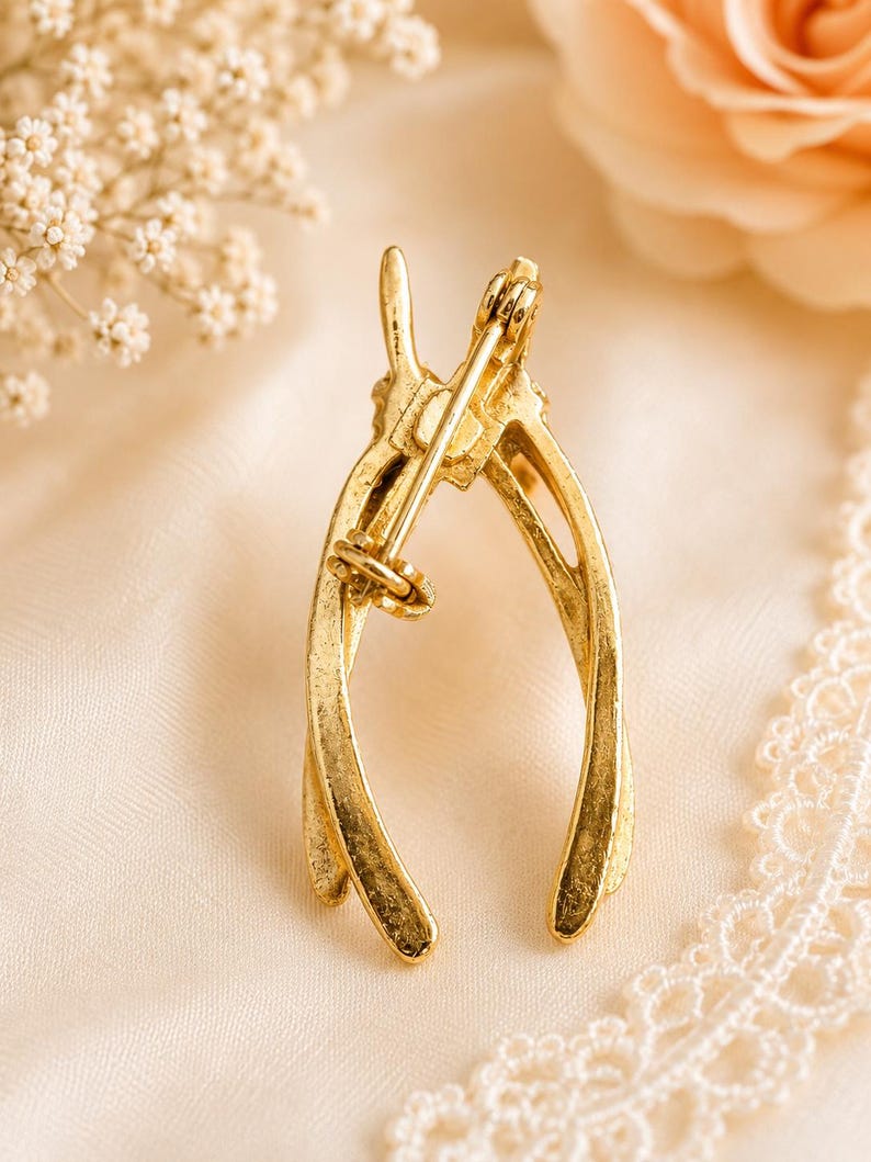 Puede incluir: Un broche de horquilla dorado, visto desde atr&aacute;s, con un cierre de alfiler. El broche est&aacute; hecho de un material met&aacute;lico brillante. El fondo es una tela suave de color blanquecino con un acento floral.