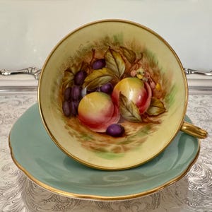 May include: A vintage teacup and saucer set. The teacup interior features a painted still life of peaches and grapes. The cup and saucer are light green with gold trim. The teacup has a gold handle.