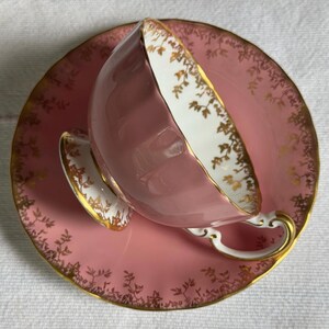 Peut inclure: Un service &agrave; th&eacute; rose compos&eacute; d'une tasse et d'une soucoupe. La tasse et la soucoupe sont orn&eacute;es de bordures dor&eacute;es et de motifs floraux. L'int&eacute;rieur de la tasse est blanc avec des bordures dor&eacute;es.