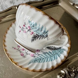 Puede incluir: Juego de taza y platillo blancos con borde dorado y dise&ntilde;o floral. La taza y el platillo tienen un borde festoneado y est&aacute;n decorados con ilustraciones bot&aacute;nicas rosas y azules. El juego est&aacute; colocado sobre una bandeja plateada.
