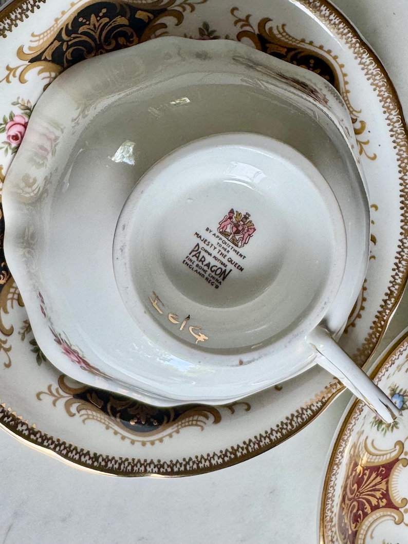 May include: A close-up of a white teacup and saucer set with gold trim and floral accents. The teacup is upside down, revealing the Paragon England mark. The set has a vintage aesthetic.