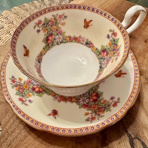 May include: A white teacup and saucer with a floral and butterfly design. The cup and saucer have a gold trim and a scalloped edge.