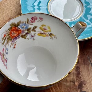 May include: A white teacup with a gold rim and floral pattern. The teacup is sitting on a turquoise saucer with a gold rim.