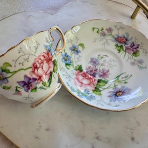 Pu&ograve; includere: Un set vintage di tazza e piattino da t&egrave; con un motivo floreale. La tazza e il piattino bianchi hanno bordi dorati e presentano fiori rosa, viola e blu. Il set &egrave; posto su un vassoio a forma di cuore.