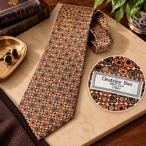 May include: A brown silk necktie featuring a repeating geometric floral pattern in shades of tan, black, and orange. A white fabric label is stitched to the back, reading Geatrice Dini, 100% Silk, Italy, in black text.