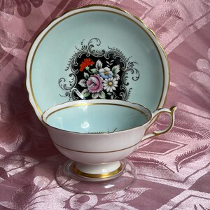 May include: A porcelain teacup and saucer set featuring a pale blue interior and white exterior with gold trim. The saucer displays a central black cartouche containing a colorful floral bouquet of pink, white, and orange flowers with green leaves.