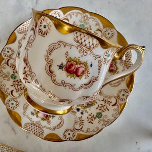 K&ouml;nnte beinhalten: Ein Vintage-Teebecher- und Untertassen-Set. Die wei&szlig;e Porzellantasse und Untertasse weisen ein Blumendesign mit roten Rosen und goldenen Verzierungen auf. Die Tasse hat einen gebogenen Henkel und steht auf einer passenden Untertasse mit gewelltem Rand.