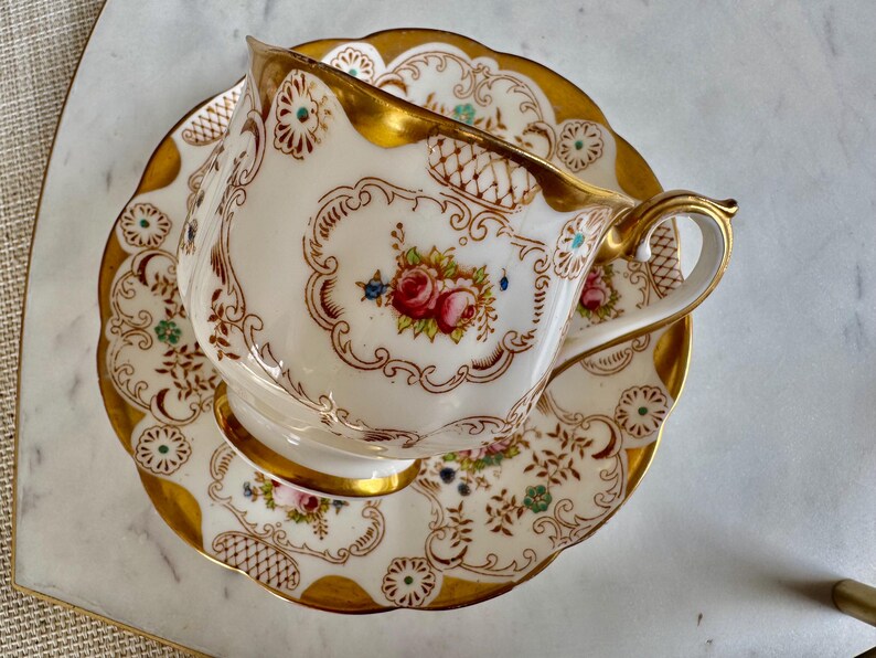K&ouml;nnte beinhalten: Ein Vintage-Teebecher- und Untertassen-Set. Die wei&szlig;e Porzellantasse und Untertasse weisen ein Blumendesign mit roten Rosen und goldenen Verzierungen auf. Die Tasse hat einen gebogenen Henkel und steht auf einer passenden Untertasse mit gewelltem Rand.