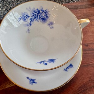 May include: A white teacup and saucer set with a blue floral pattern. The cup and saucer have a gold trim around the edges.