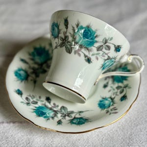 Peut inclure: Service &agrave; th&eacute; blanc avec une tasse et une soucoupe orn&eacute;es de roses turquoise et de bordures dor&eacute;es. La tasse a une anse d&eacute;licate et la soucoupe a un bord festonn&eacute;. Le motif floral comprend des roses, des boutons et des feuilles gris-vert.