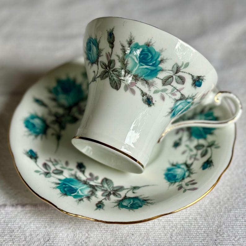 Peut inclure: Service &agrave; th&eacute; blanc avec une tasse et une soucoupe orn&eacute;es de roses turquoise et de bordures dor&eacute;es. La tasse a une anse d&eacute;licate et la soucoupe a un bord festonn&eacute;. Le motif floral comprend des roses, des boutons et des feuilles gris-vert.