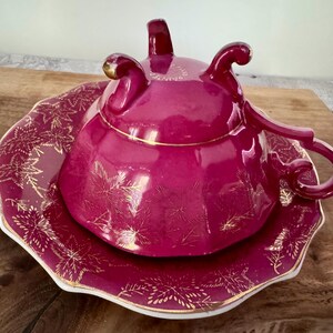 May include: A magenta-colored teacup and saucer set. The teacup has a lid and a curved handle. The saucer and teacup are decorated with gold leaf designs. The edges of the saucer and teacup are trimmed with gold. The set is on a wooden surface.