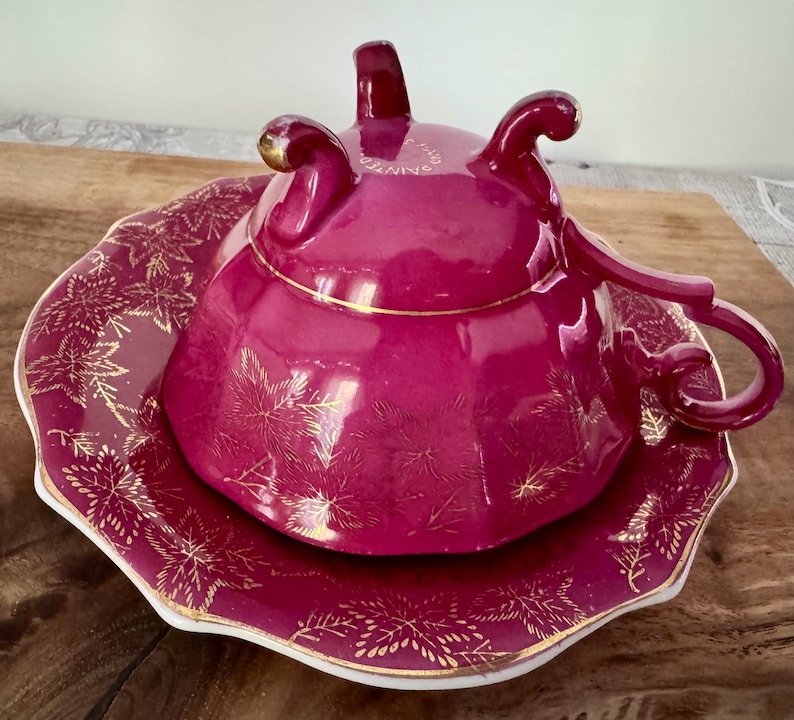 May include: A magenta-colored teacup and saucer set. The teacup has a lid and a curved handle. The saucer and teacup are decorated with gold leaf designs. The edges of the saucer and teacup are trimmed with gold. The set is on a wooden surface.