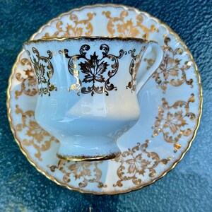 May include: A white porcelain teacup and matching saucer set featuring a decorative gold leaf pattern. The cup has a scalloped rim and a handle, while the saucer has a matching scalloped edge. Both pieces are adorned with intricate gold filigree.