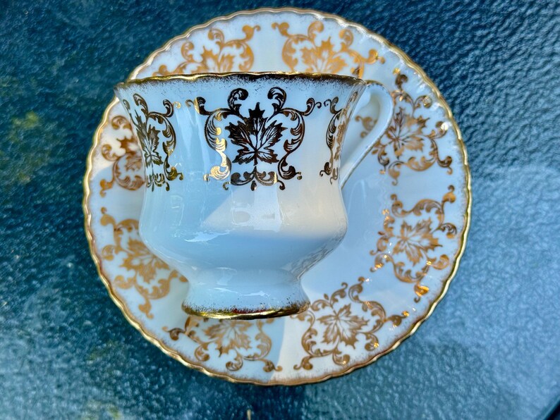 May include: A white porcelain teacup and matching saucer set featuring a decorative gold leaf pattern. The cup has a scalloped rim and a handle, while the saucer has a matching scalloped edge. Both pieces are adorned with intricate gold filigree.