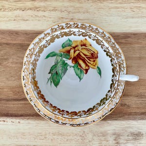 Puede incluir: Juego de taza y platillo blancos con borde floral dorado y una gran rosa amarilla y roja en el centro. La taza tiene un asa blanca. El juego est&aacute; sobre una superficie de madera.