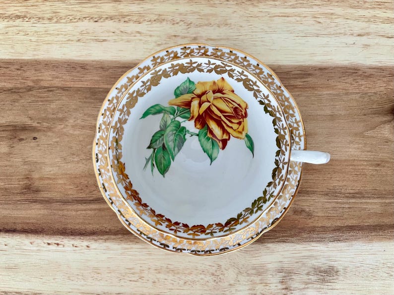 Puede incluir: Juego de taza y platillo blancos con borde floral dorado y una gran rosa amarilla y roja en el centro. La taza tiene un asa blanca. El juego est&aacute; sobre una superficie de madera.