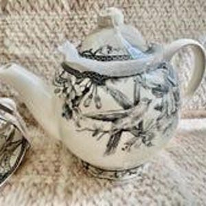 May include: A cream-colored ceramic teapot featuring a detailed black and white botanical illustration of birds perched on flowering branches. The teapot has a matching lid with a knob handle, a curved spout, and a sturdy handle on the side.
