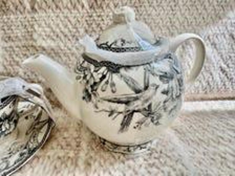 May include: A cream-colored ceramic teapot featuring a detailed black and white botanical illustration of birds perched on flowering branches. The teapot has a matching lid with a knob handle, a curved spout, and a sturdy handle on the side.