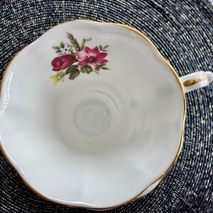 Puede incluir: Una taza de t&eacute; blanca con un borde dorado y un dise&ntilde;o floral con rosas rosas y rojas. La taza de t&eacute; tiene un borde festoneado y un asa.
